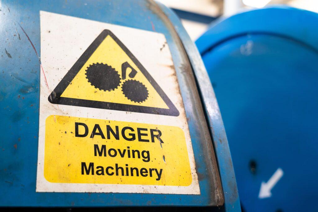 Warnschild auf blauer Maschine mit Aufschrift "DANGER Moving Machinery" und grafischer Darstellung von Zahnrädern, illustriert die Wichtigkeit klarer Sicherheitshinweise und Typenschilder auf Industrieanlagen.