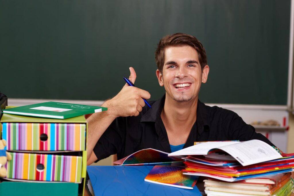 Fröhlicher junger Lehrer im Klassenzimmer zeigt Daumen hoch, umgeben von bunten Ordnern, symbolisiert die Bedeutung einer soliden Krankenversicherung im Referendariat.