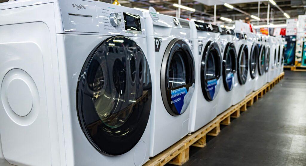 Reihe neuer Waschmaschinen in einem Elektronikmarkt, ausgestellt auf Paletten, mit deutlich sichtbaren Typenschildern, die wichtige Informationen wie Marke, Modell und Energieeffizienz anzeigen.