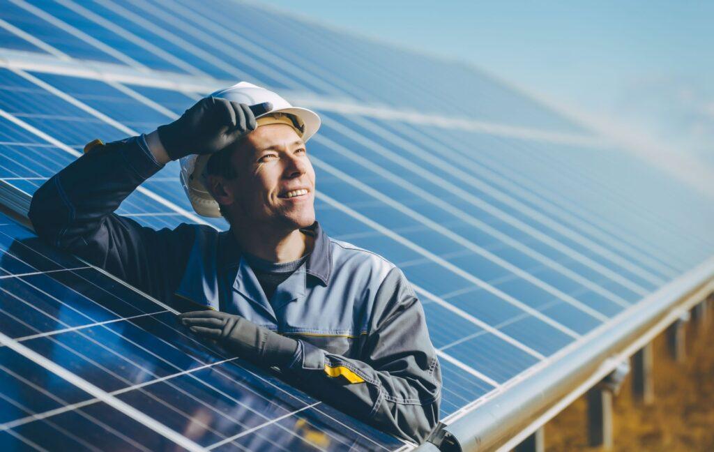 Ein lachender Techniker steht vor einer großen Photovoltaikanlage und überprüft die Solarmodule bei sonnigem Wetter.