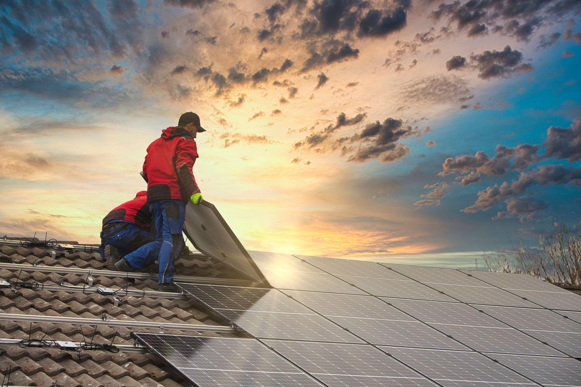 Zwei Techniker installieren Solarmodule auf einem Dach bei Sonnenuntergang. Die Abendsonne spiegelt sich auf den Photovoltaik-Paneelen wider und sorgt für eine stimmungsvolle Atmosphäre.