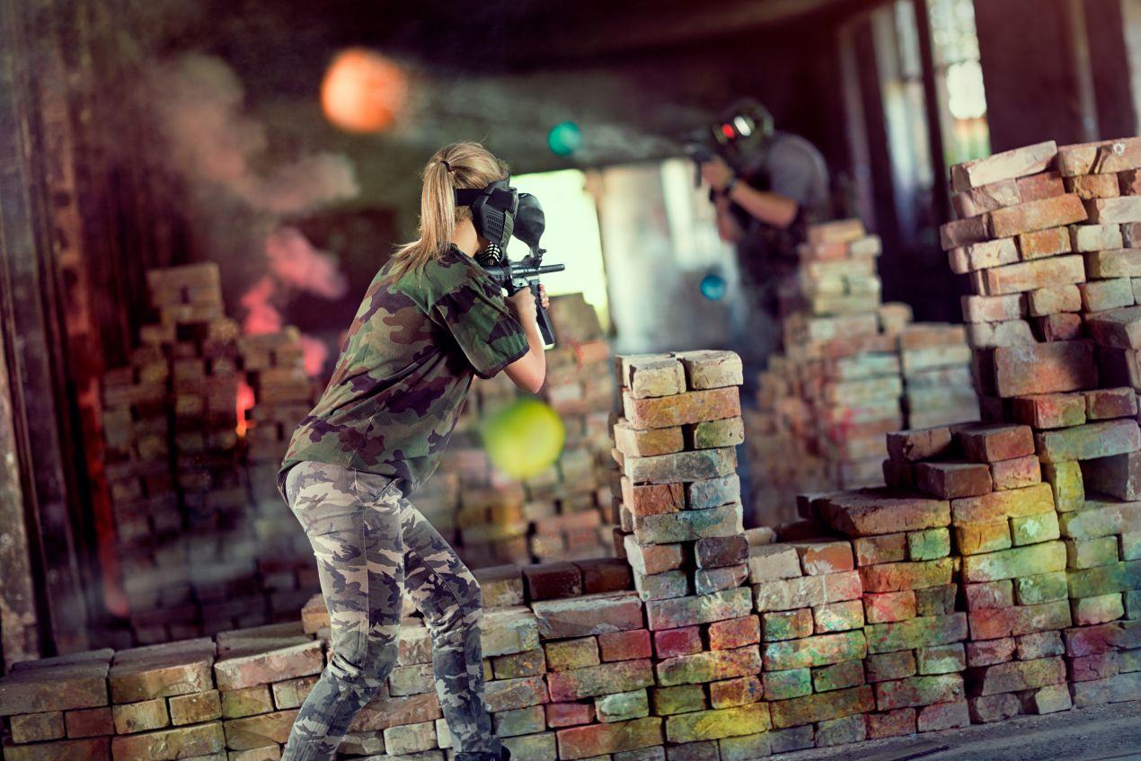 Paintball-Spielerin hinter Mauer aus Ziegelsteinen in Indoor-Anlage zielt mit Druckluftwaffe – Fokus auf Struktur und Spielfeldsicherheit