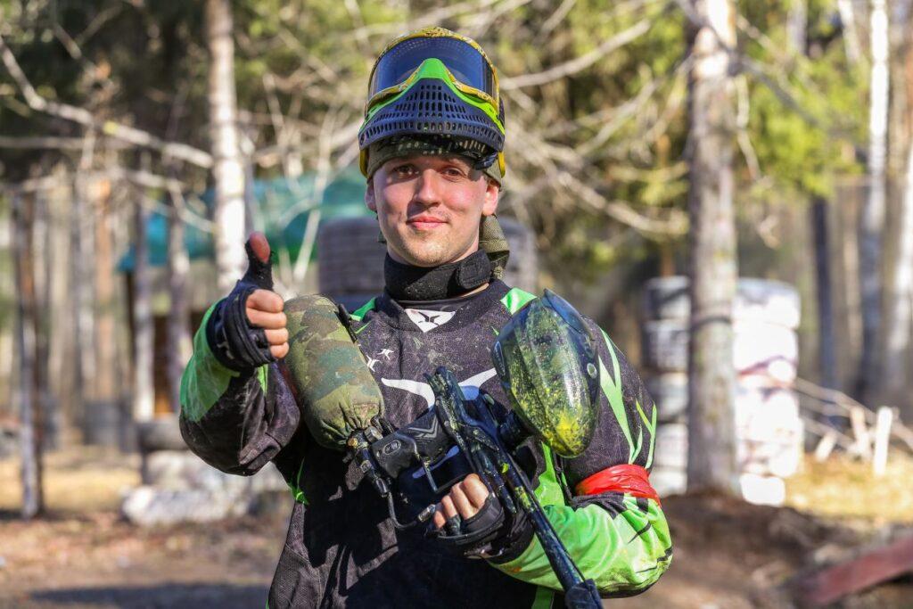Paintball-Spieler mit kompletter Schutzkleidung haelt Druckluftwaffe und zeigt Daumen hoch – Symbol fuer sichere Techniknutzung im Freizeitbereich