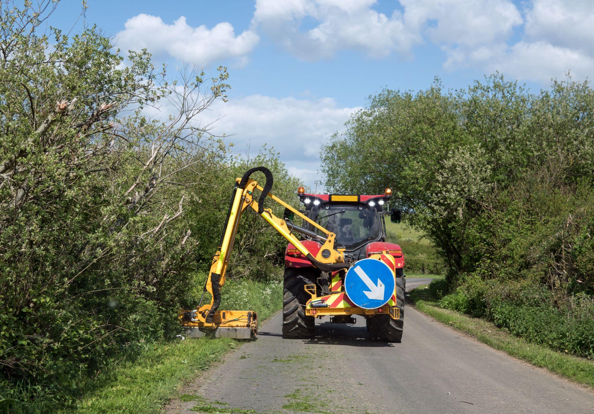 Traktor mit hydraulischem Ausleger schneidet Randbewuchs an schmaler Landstrasse mit gelbem Boeschungsmulcher